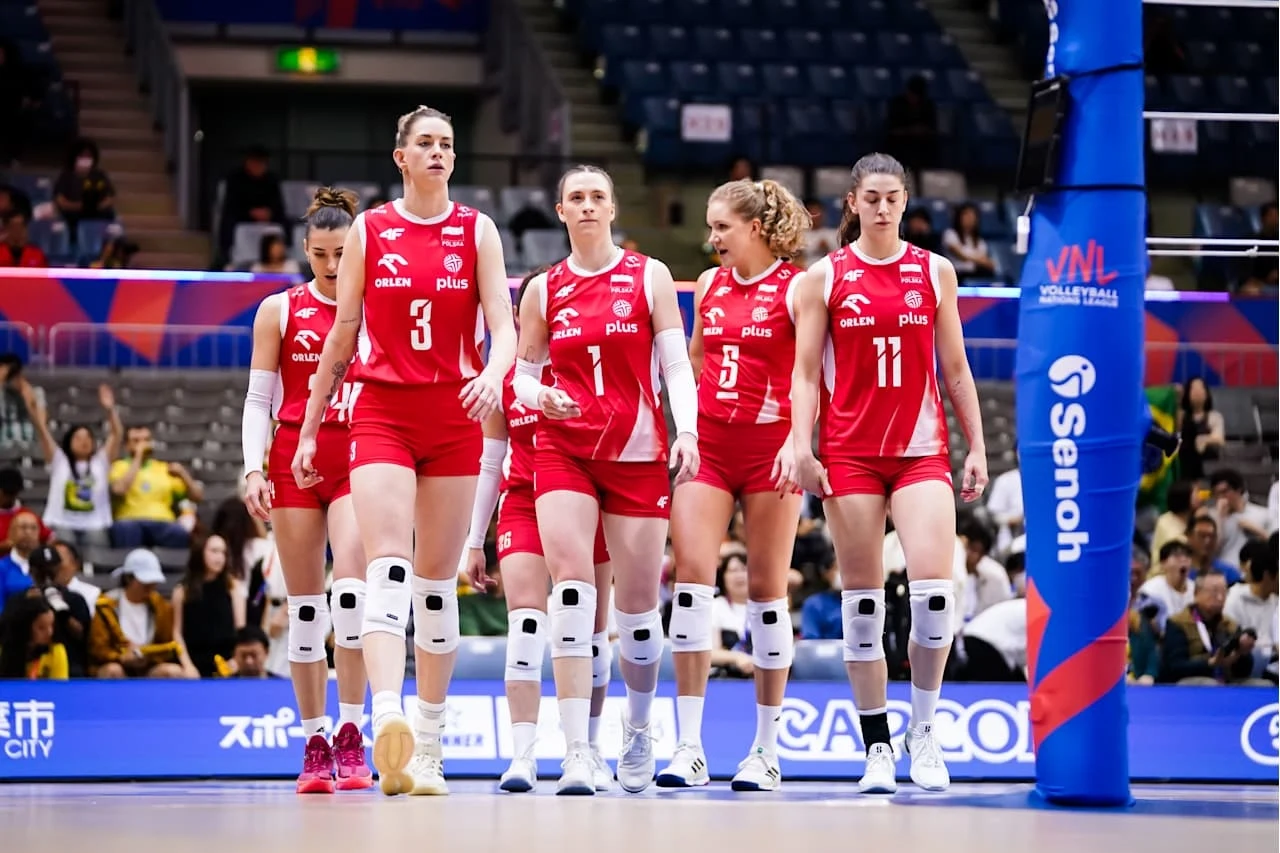 Grupa siatkarek w czerwonych strojach, wyposażonych w ochraniacze na kolana, wchodzi na boisko podczas meczu halowego, wokół widoczni kibice na trybunach oraz sportowy sprzęt i elementy wyposażenia hali.