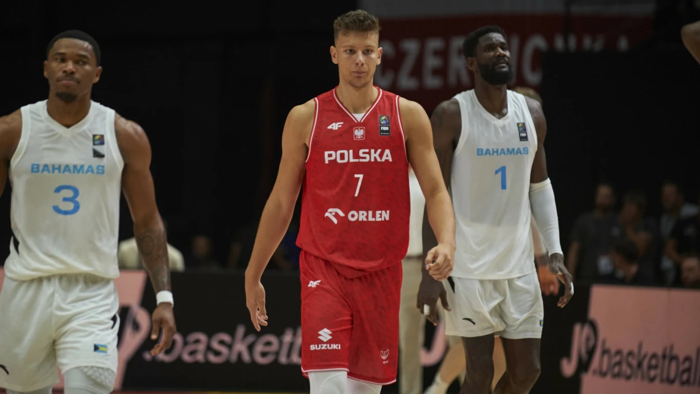 Zawodnik polskiej reprezentacji koszykówki w czerwonym stroju z numerem 7 otoczony przez dwóch zawodników drużyny Bahamów w białych strojach podczas meczu na parkiecie, w tle widoczna publiczność i fragmenty reklam. Zawodnik polskiej reprezentacji koszykówki w czerwonym stroju z numerem 7 otoczony przez dwóch zawodników drużyny Bahamów w białych strojach podczas meczu na parkiecie, w tle widoczna publiczność i fragmenty reklam.