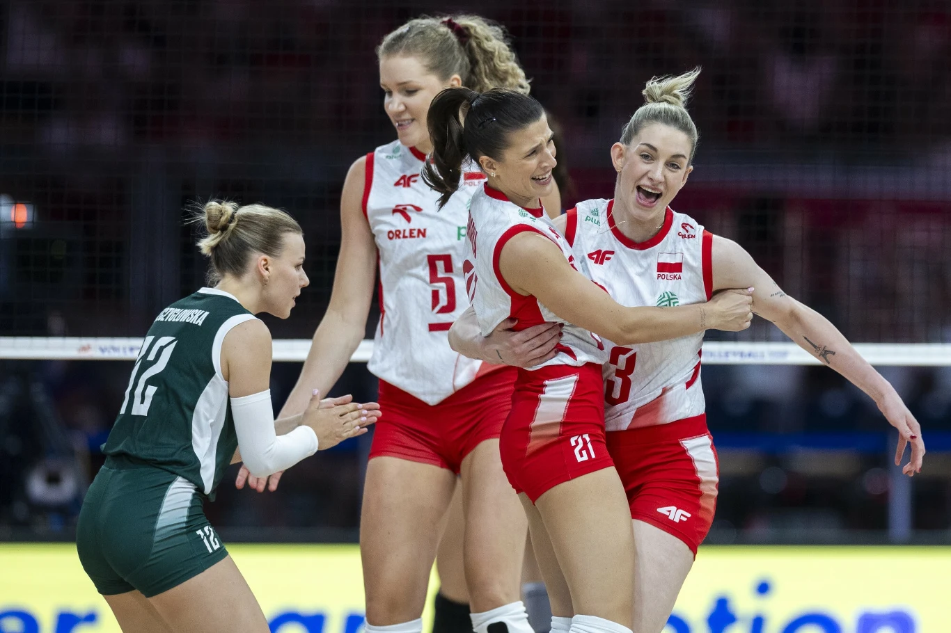 Grupa siatkarek w biało-czerwonych strojach świętuje zdobyty punkt, na pierwszym planie radość oraz gesty triumfu, obok zawodniczka drużyny przeciwnej w zielonym stroju.