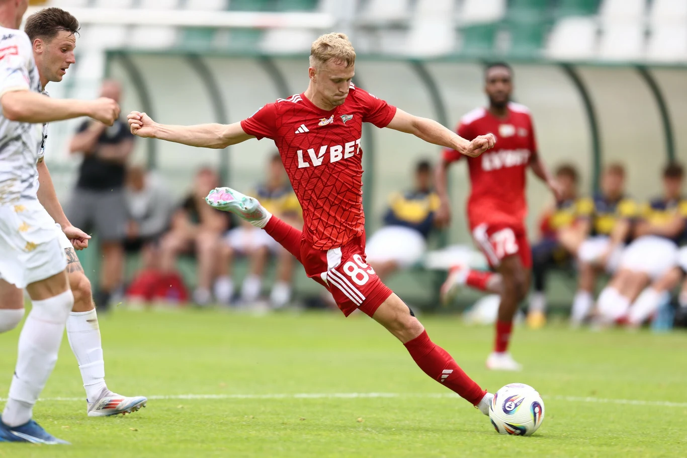 Piłkarz w czerwonym stroju kopie piłkę podczas meczu piłki nożnej, w tle widoczni są inni zawodnicy oraz kibice na trybunach.