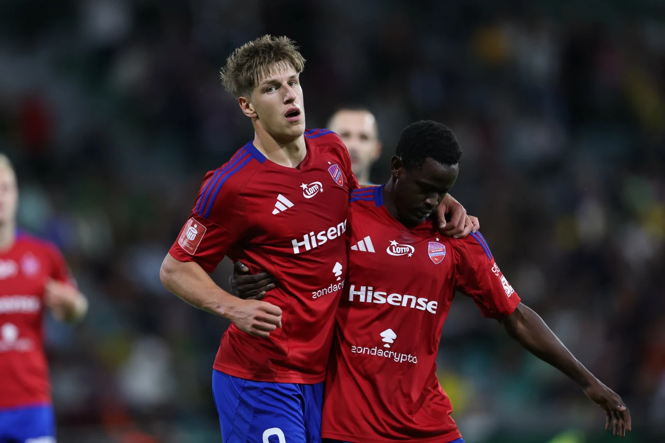 Dwóch piłkarzy w czerwonych koszulkach z logotypami sponsorów, jeden obejmuje drugiego ramieniem, na rozmytym tle widoczny fragment boiska i postacie innych zawodników.