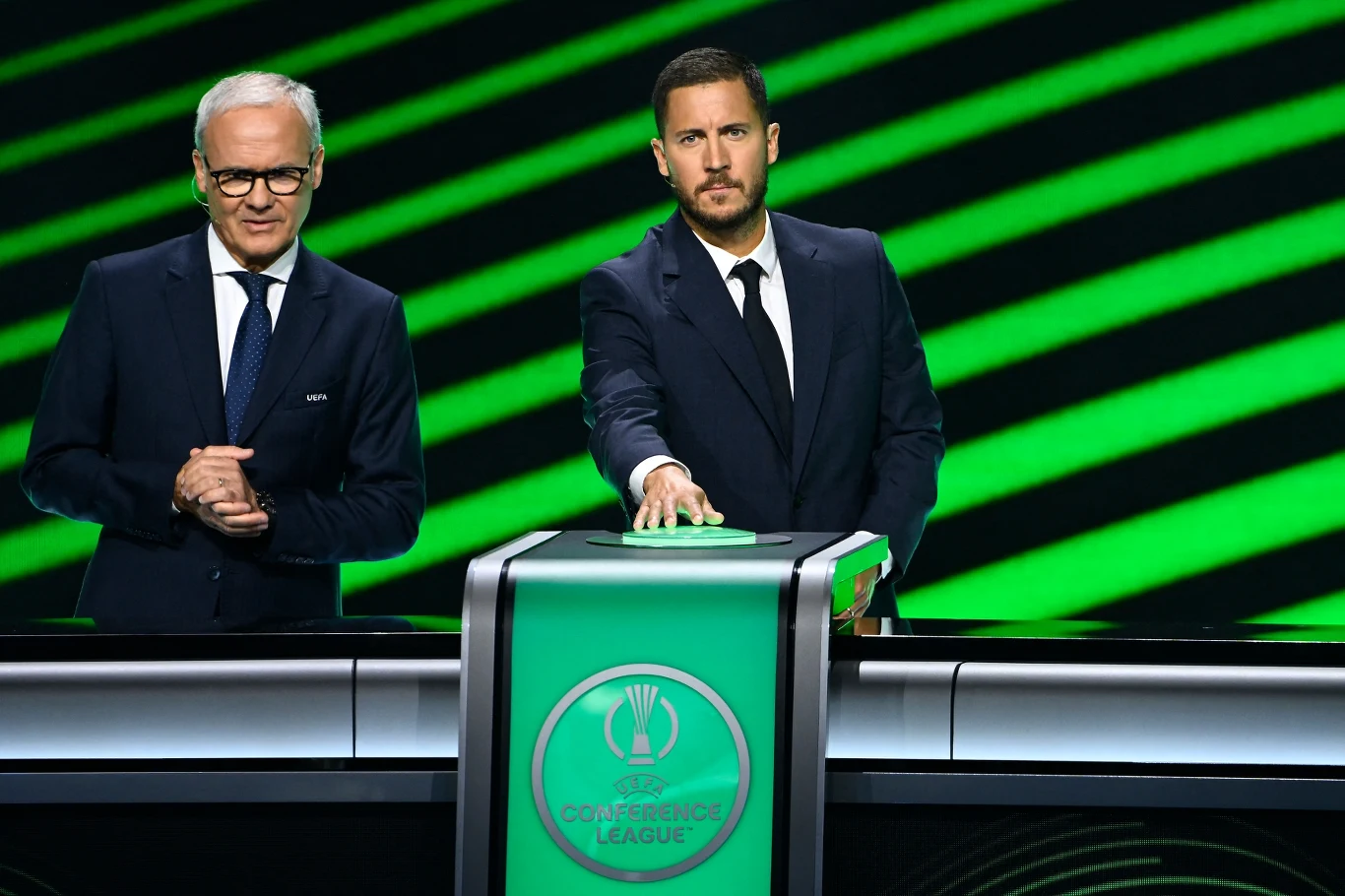 Dwóch mężczyzn ubranych w eleganckie garnitury stoi przy podium z logo UEFA Conference League, jeden z nich naciska przycisk, tło w zielone ukośne linie. Dwóch mężczyzn ubranych w eleganckie garnitury stoi przy podium z logo UEFA Conference League, jeden z nich naciska przycisk, tło w zielone ukośne linie.