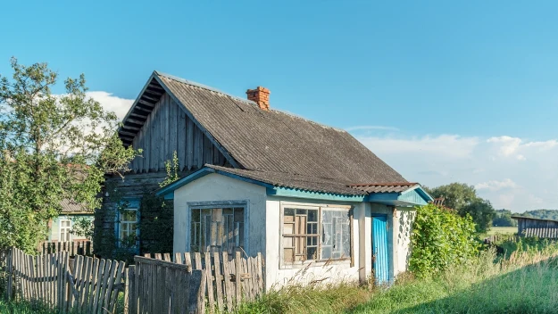 Drewniany, stary dom z białą przybudówką i niebieskimi akcentami, otoczony drewnianym rozwalonym płotem, w letniej, wiejskiej scenerii z bujną zielenią dookoła.