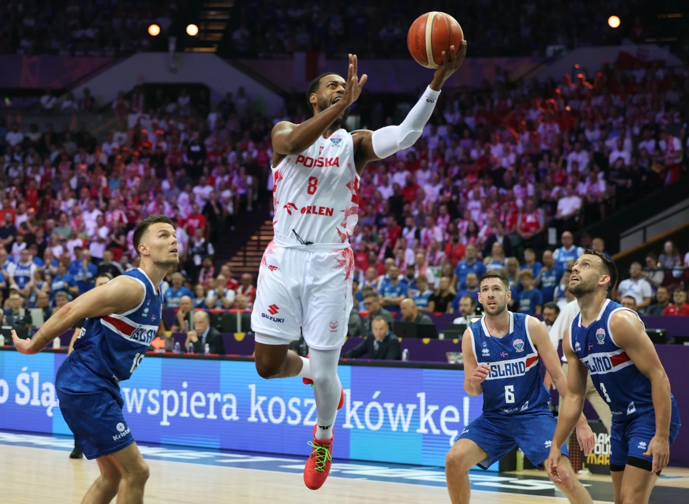 Zawodnik reprezentacji Polski wykonuje dynamiczny rzut do kosza podczas meczu koszykówki z drużyną Islandii. W tle widoczna pełna trybuna kibiców oraz trzech zawodników drużyny przeciwnej próbujących uniemożliwić zdobycie punktów.