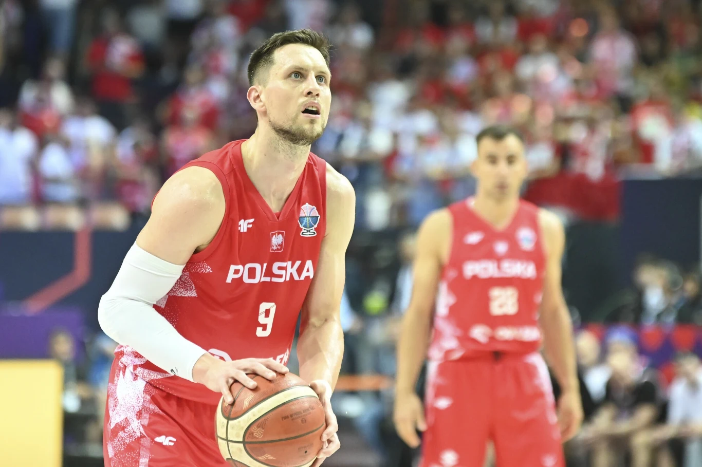 Koszykarz drużyny narodowej Polski w czerwonym stroju przygotowuje się do wykonania rzutu wolnego, w tle widoczny drugi zawodnik w podobnym stroju oraz rozmyta publiczność. Koszykarz drużyny narodowej Polski w czerwonym stroju przygotowuje się do wykonania rzutu wolnego, w tle widoczny drugi zawodnik w podobnym stroju oraz rozmyta publiczność.