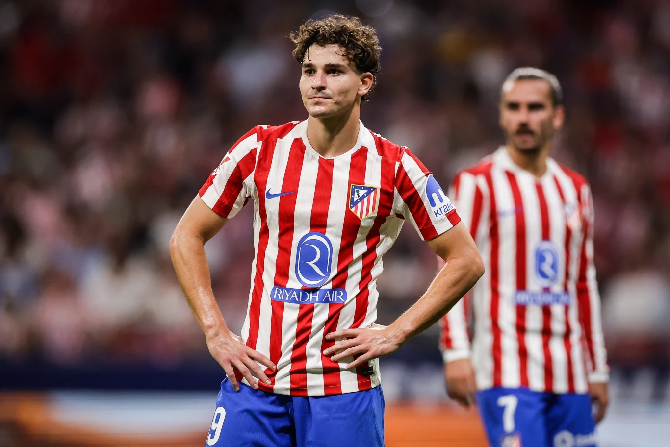 Dwóch piłkarzy w czerwono-białych pasiastych koszulkach oraz niebieskich spodenkach stoi na boisku podczas meczu, w tle rozmyte postaci oraz kibice na trybunach.