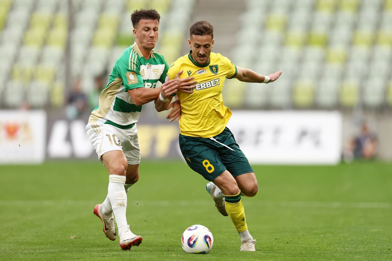 Dwóch piłkarzy w trakcie dynamicznej walki o piłkę na boisku podczas meczu, jeden ubrany w zielono-biały strój, drugi w żółto-zielony, w tle trybuny stadionu.