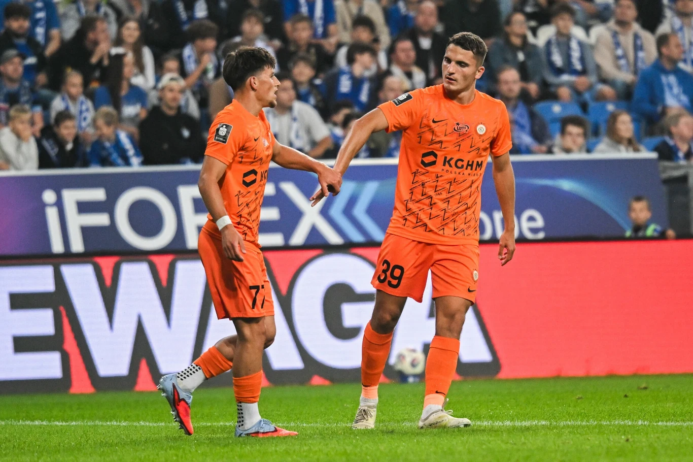 Dwóch piłkarzy w pomarańczowych strojach drużyny KGHM Zagłębie Lubin stoi na murawie stadionu, za nimi widoczna publiczność i reklamy na bandach.