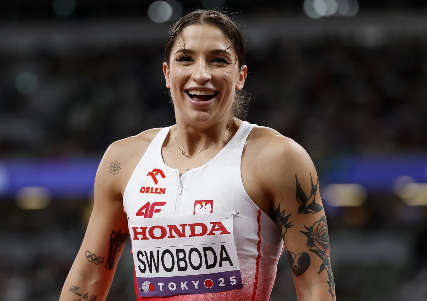 Uśmiechnięta lekkoatletka w stroju sportowym, z widocznymi tatuażami na rękach, z numerem startowym, na tle stadionu lekkoatletycznego w trakcie zawodów.