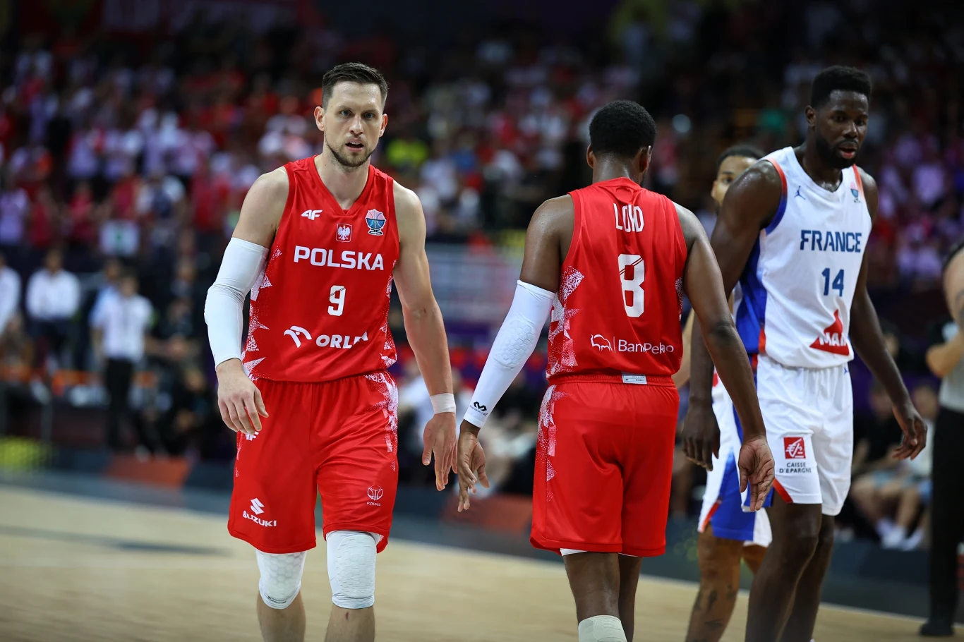 Dwóch koszykarzy w czerwonych strojach polskiej reprezentacji na pierwszym planie, po lewej stronie zawodnik numer 9, obok niego zawodnik numer 8. W tle widoczny koszykarz drużyny Francji w białym stroju i rozgrywka na parkiecie sportowej hali.