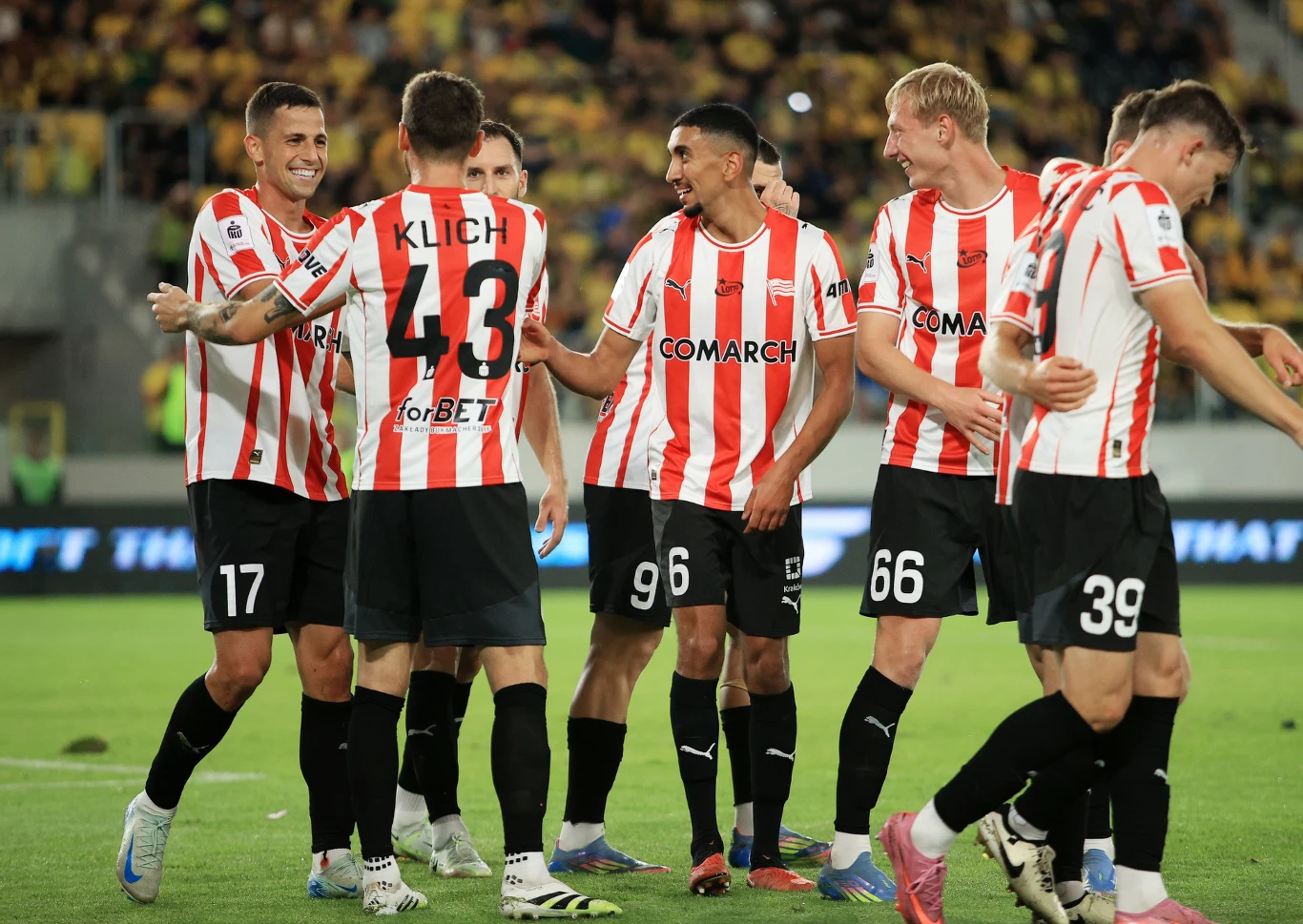 Grupa piłkarzy w czerwono-białych pasiastych koszulkach świętuje na boisku, widoczne uśmiechy i wzajemne gratulacje, w tle kibice na trybunach.