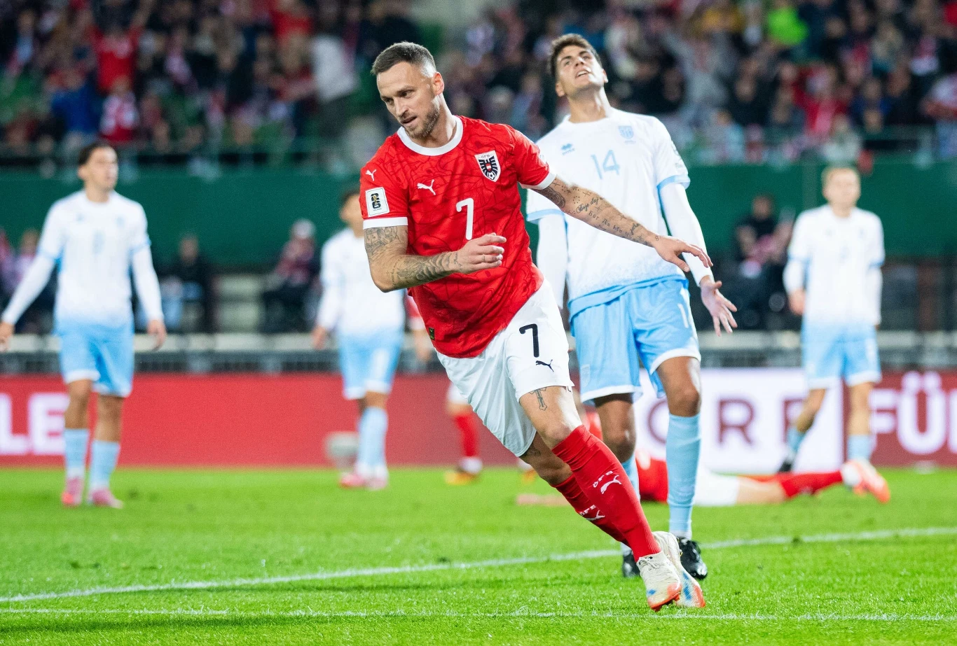 Piłkarz w czerwonej koszulce z numerem 7 celebruje strzelenie gola podczas meczu na stadionie, w tle zawodnicy drużyny przeciwnej w jasnoniebieskich strojach oraz dopingujący kibice.