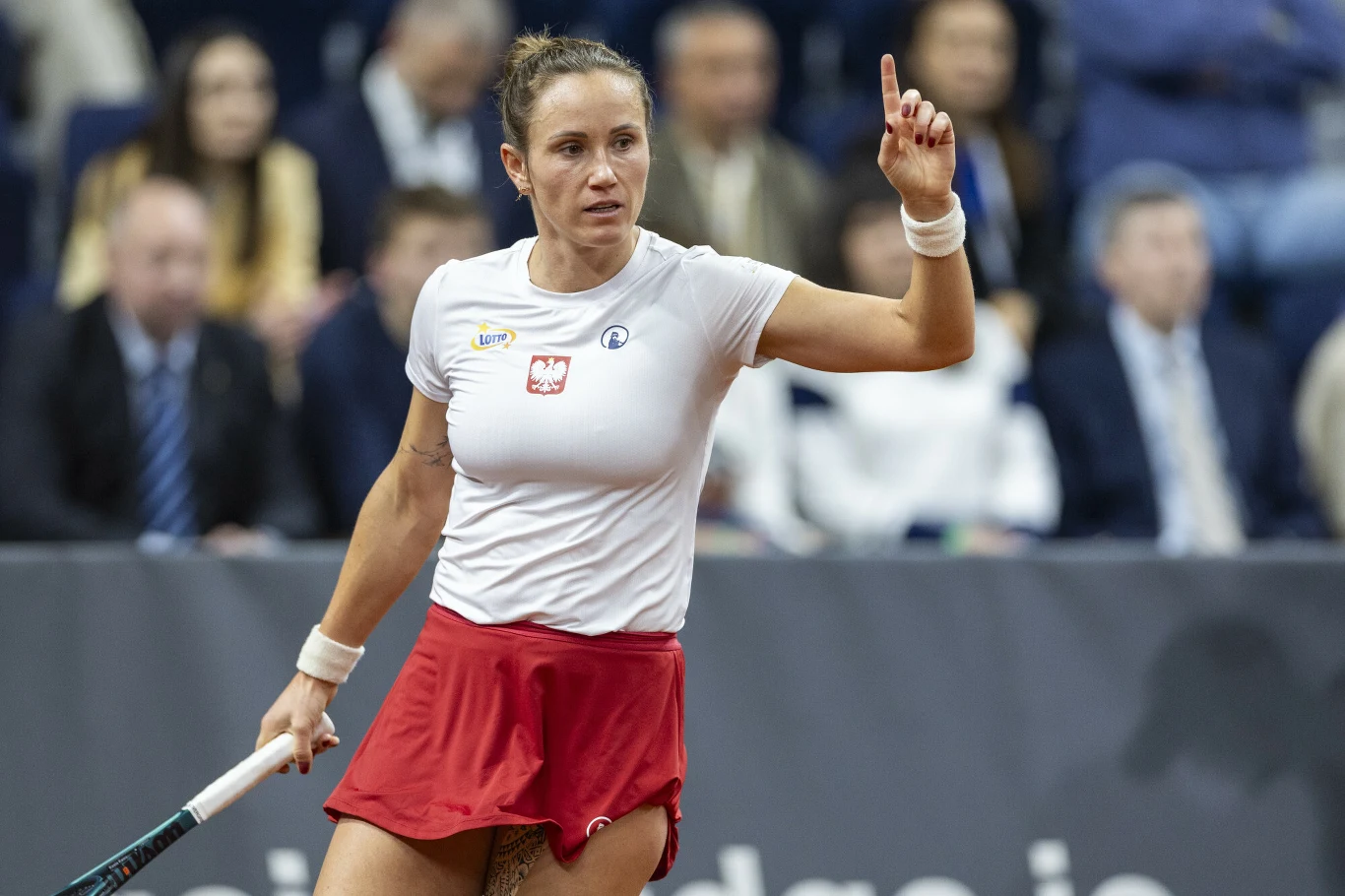 Tenisistka w czerwonej spódniczce i białej koszulce z godłem Polski podnosi rękę z wyciągniętym palcem wskazującym, trzymając rakietę, na tle rozmytej publiczności.