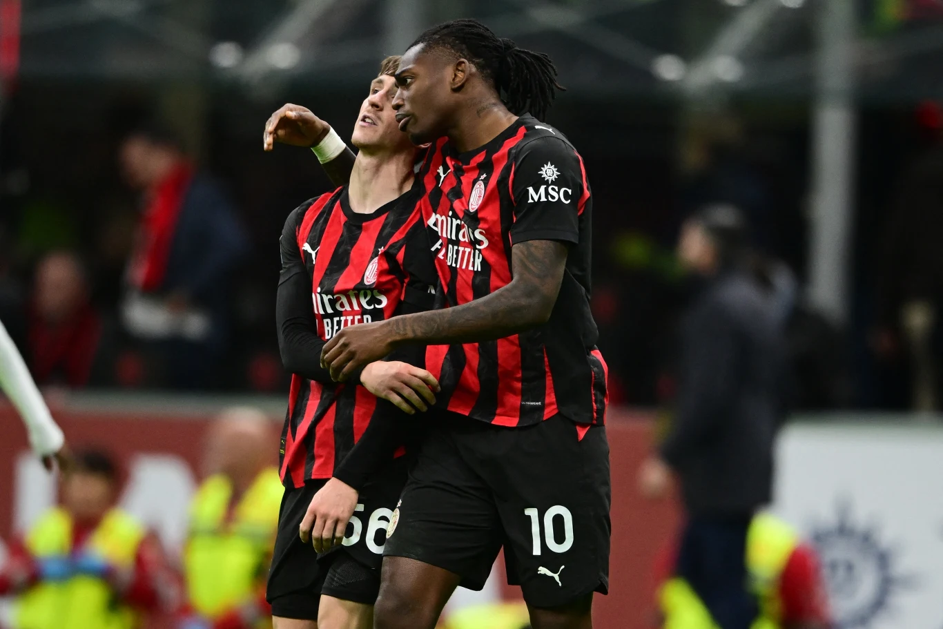 Dwóch piłkarzy drużyny AC Milan w czarno-czerwonych strojach ściska się na boisku, jeden obejmuje drugiego ramieniem, w tle rozmyty obraz trybun z kibicami.