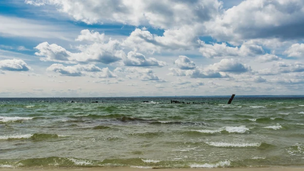 Plaża z falującą wodą i fragmentem wraku statku lub samolotu wystającym ponad powierzchnię morza, nad całością rozpościera się niebo z licznymi chmurami. Plaża z falującą wodą i fragmentem wraku statku lub samolotu wystającym ponad powierzchnię morza, nad całością rozpościera się niebo z licznymi chmurami.