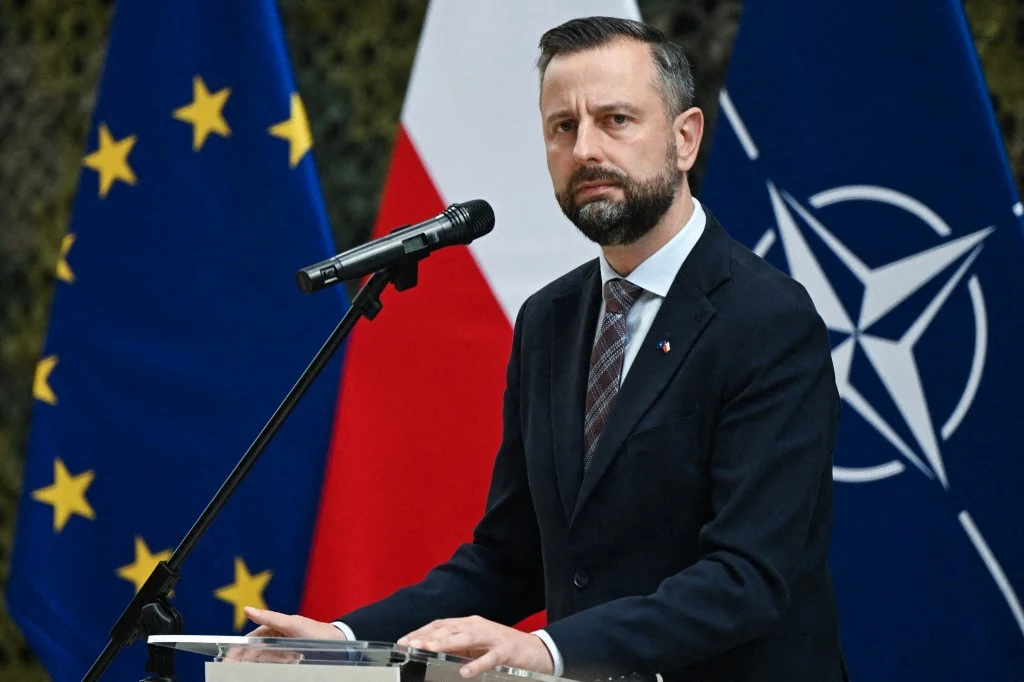 Mężczyzna w garniturze przemawia przy mównicy, za nim widoczne flagi Unii Europejskiej, Polski oraz NATO.