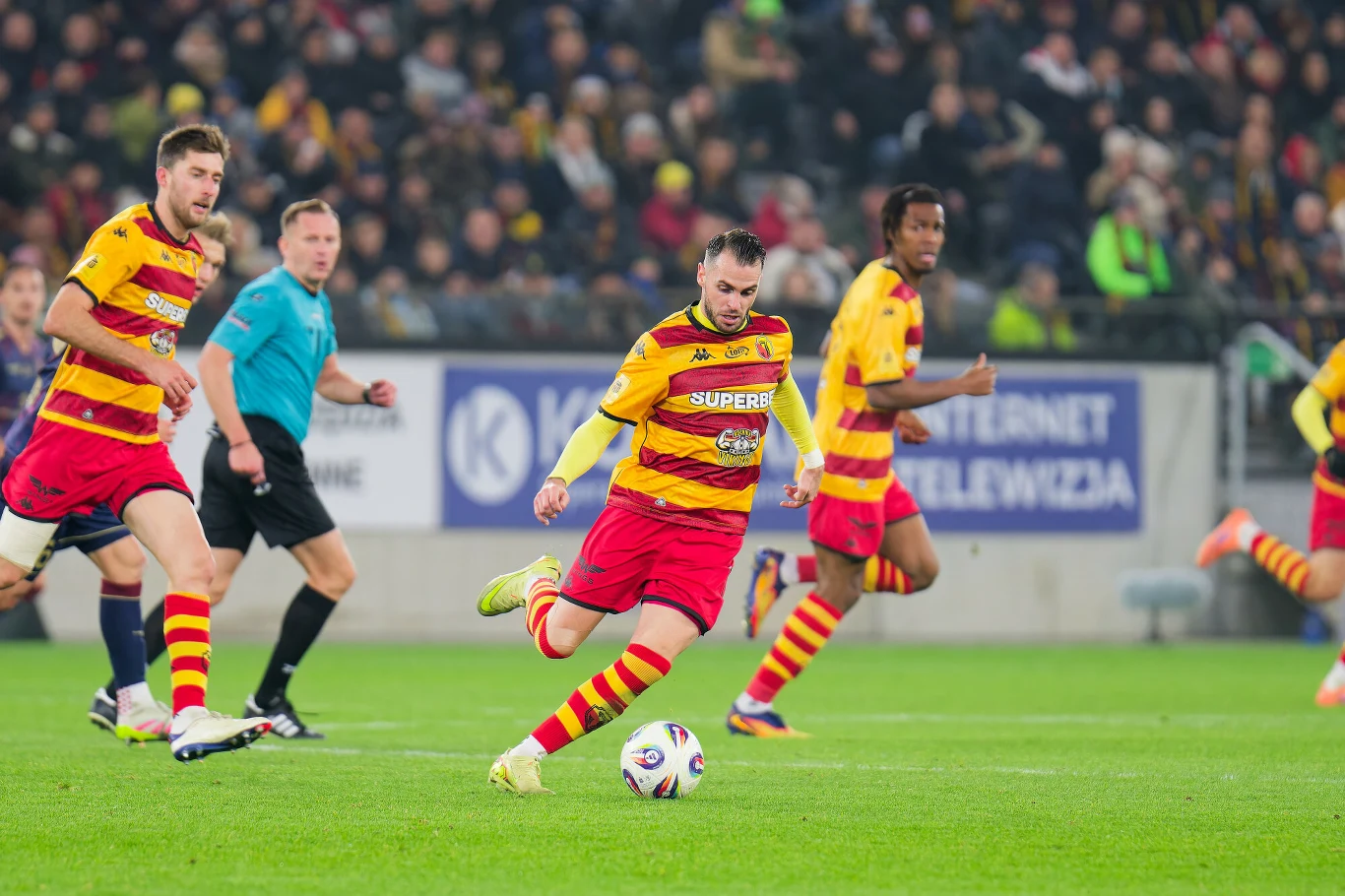 Piłkarz drużyny w żółto-czerwonych barwach w trakcie dynamicznej akcji z piłką na murawie, w tle widoczni inni zawodnicy oraz sędzia; trybuny pełne kibiców.