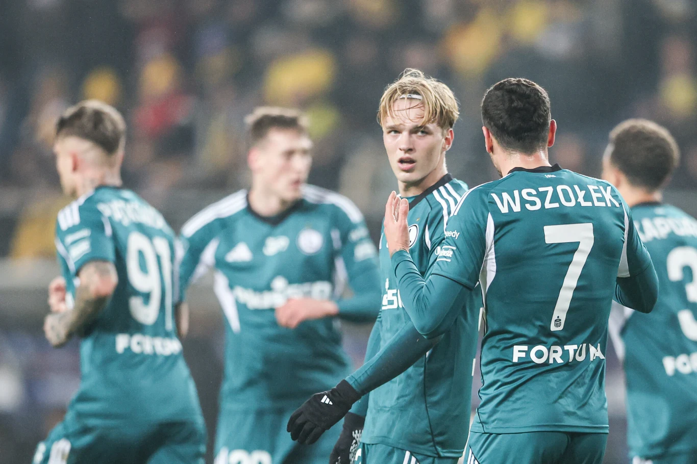 Grupa piłkarzy w zielonych strojach drużyny Śląsk Wrocław na boisku podczas meczu, jeden z nich spogląda prosto przed siebie, a drugi w koszulce z numerem 7 rozmawia z kolegą z zespołu wokół rozmyte tło trybun stadionu. Grupa piłkarzy w zielonych strojach drużyny Śląsk Wrocław na boisku podczas meczu, jeden z nich spogląda prosto przed siebie, a drugi w koszulce z numerem 7 rozmawia z kolegą z zespołu wokół rozmyte tło trybun stadionu.