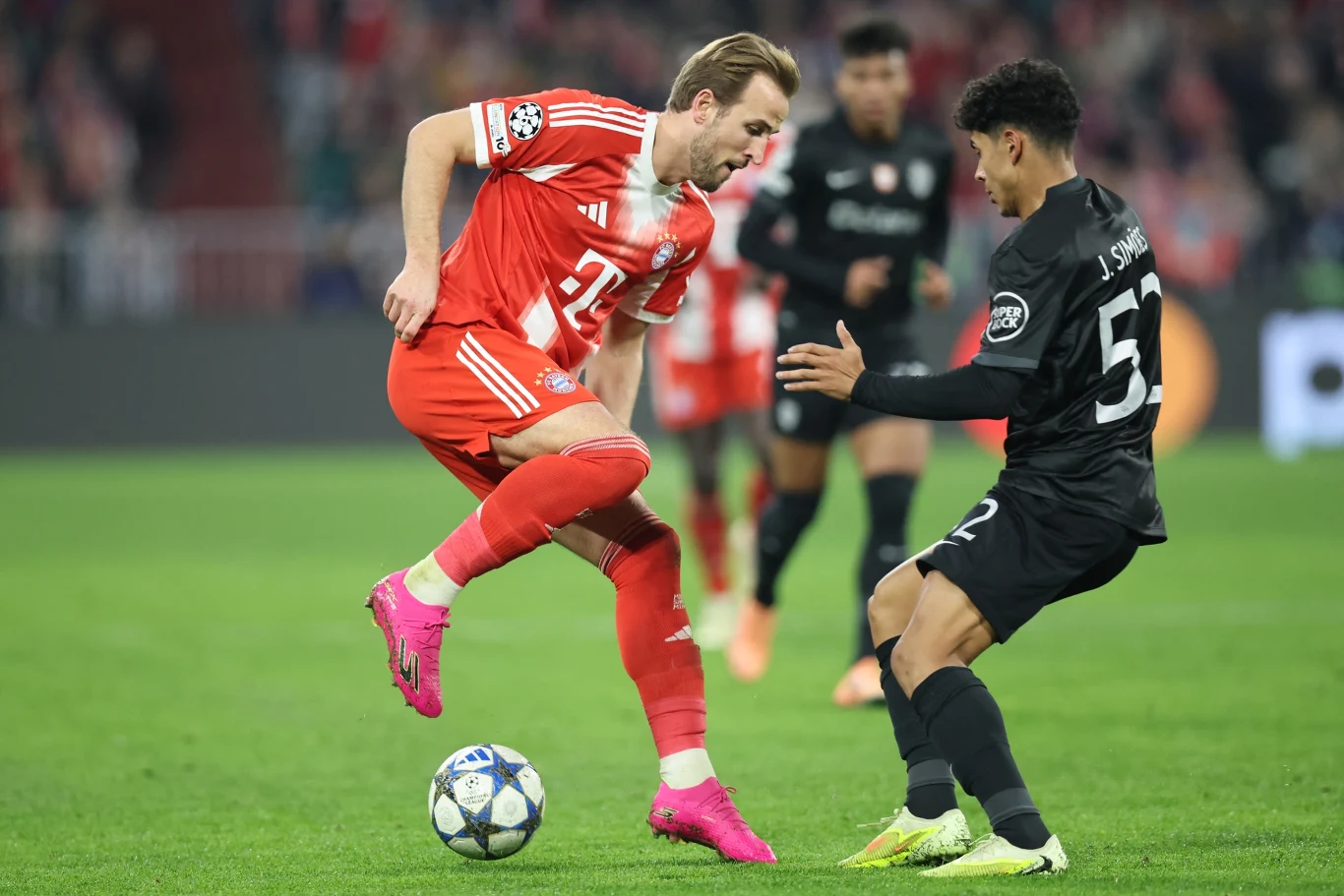 Dwóch piłkarzy walczy o piłkę podczas meczu, jeden z nich ubrany w czerwony strój, drugi w czarny, otoczeni rozmytym tłem stadionu i publicznością.