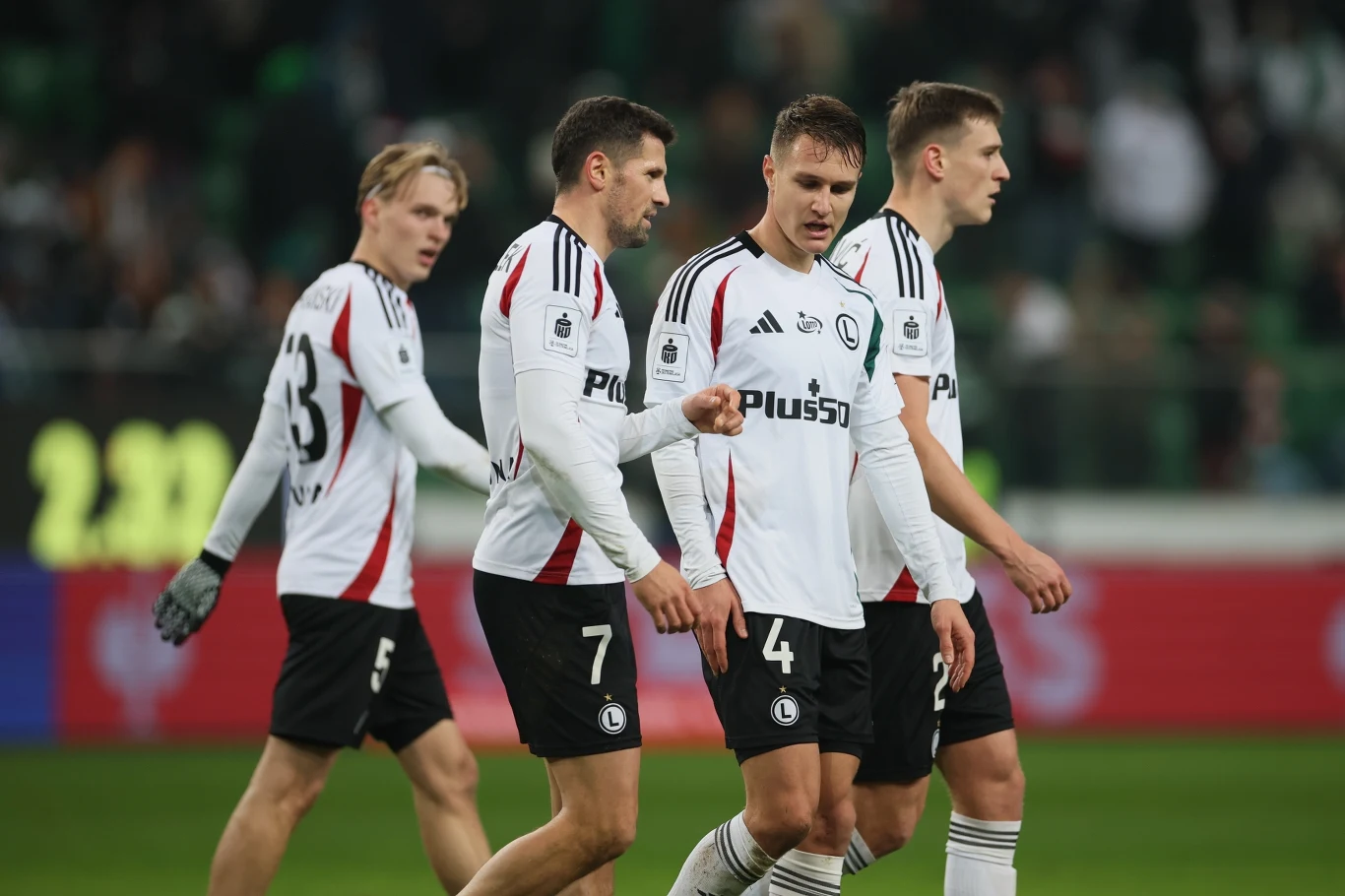 Czterech piłkarzy drużyny Legia Warszawa ubranych w biało-czarne stroje klubowe idzie razem po murawie stadionu podczas meczu piłkarskiego, w tle nieostry widok trybun i kibiców.