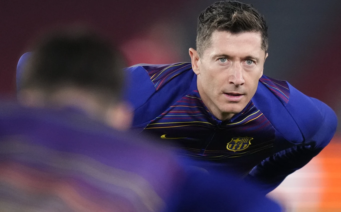 Skoncentrowany piłkarz w barwach FC Barcelony wykonuje rozgrzewkę na boisku, tło rozmyte i ciemne, w kadrze widoczna jest także sylwetka innego zawodnika z przodu.