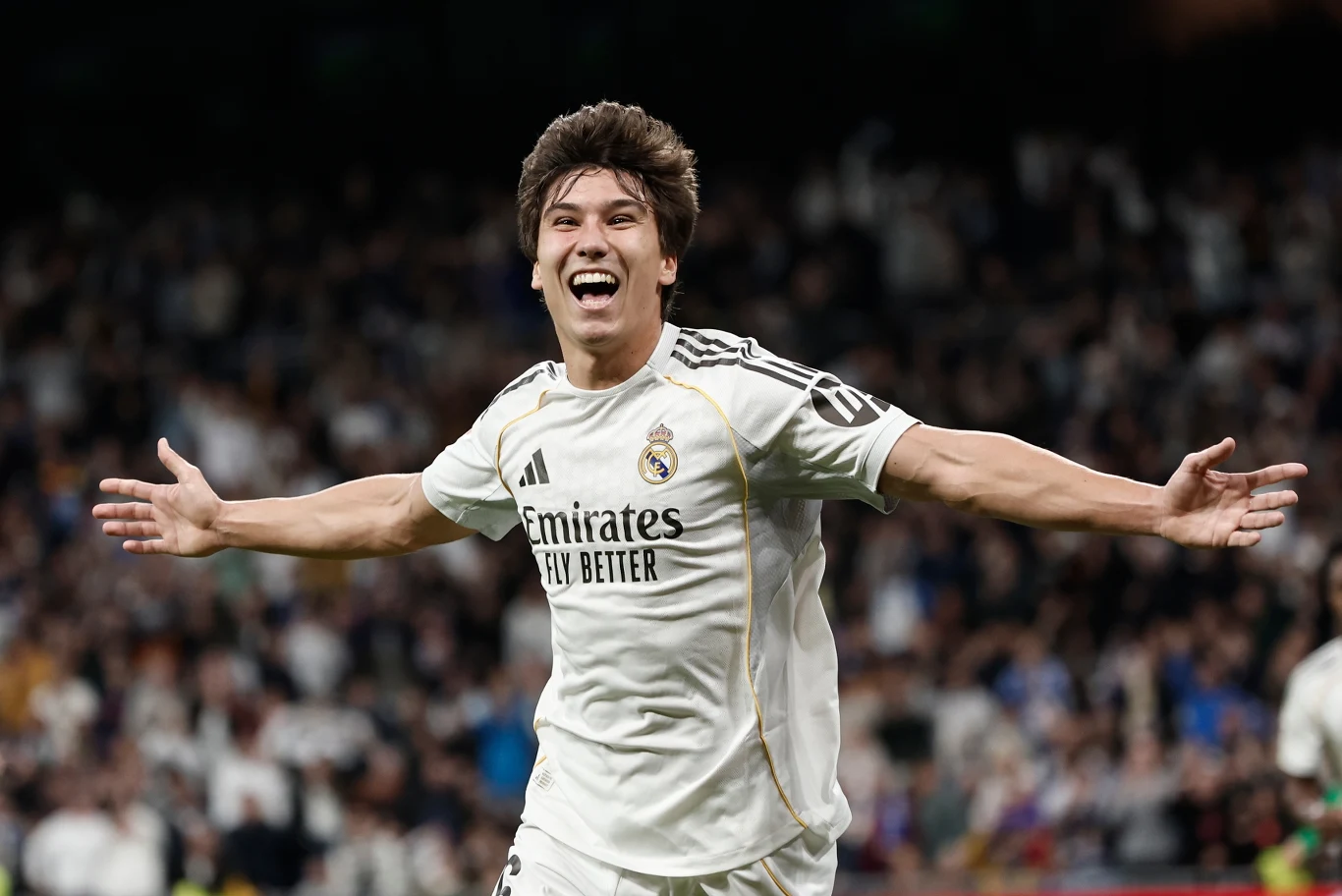 Piłkarz w białym stroju drużyny Real Madryt z szeroko rozpostartymi ramionami świętuje zdobycie gola na tle rozmytej publiczności.