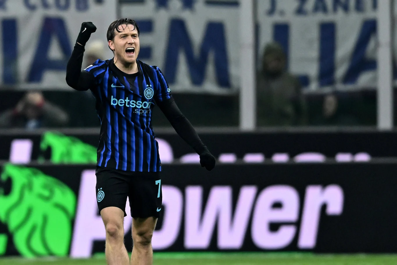 Piłkarz w czarno-niebieskiej koszulce Interu Mediolan świętuje sukces na boisku, z uniesioną ręką i wyrazem radości na twarzy. W tle bannery kibiców oraz fragment stadionu.