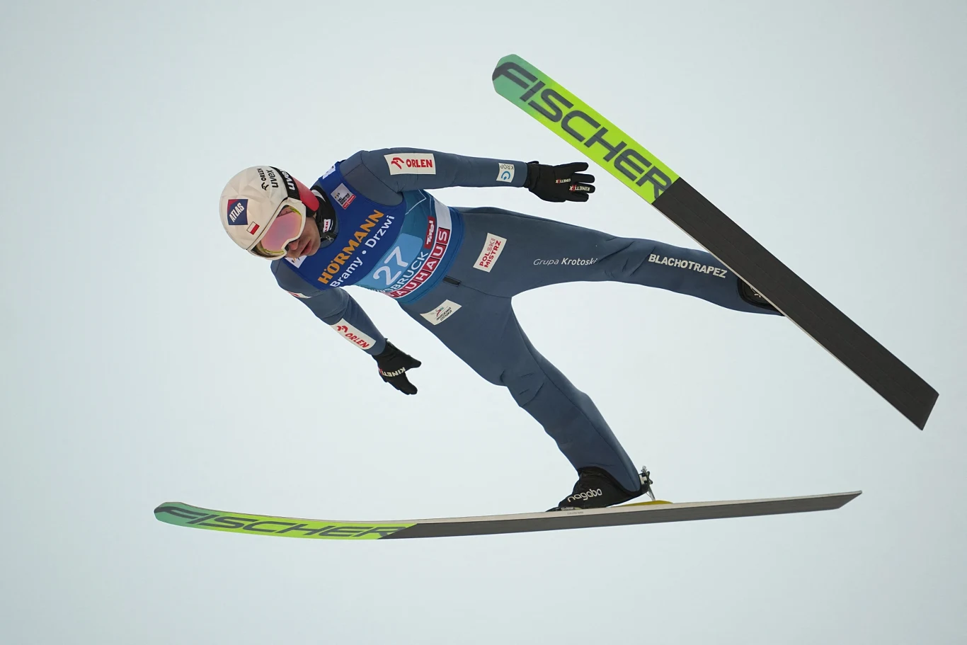 Skoczek narciarski w locie podczas zawodów na tle jasnego nieba, w pełnym sportowym ekwipunku, w pozie charakterystycznej dla skoków narciarskich z widocznymi nartami i numerem startowym.