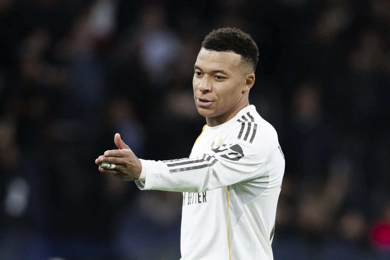 Piłkarz w białym stroju sportowym z numerem na plecach wykonuje gest obiema rękami, stojąc na stadionie podczas meczu, w tle rozmyte sylwetki widzów. Piłkarz w białym stroju sportowym z numerem na plecach wykonuje gest obiema rękami, stojąc na stadionie podczas meczu, w tle rozmyte sylwetki widzów.