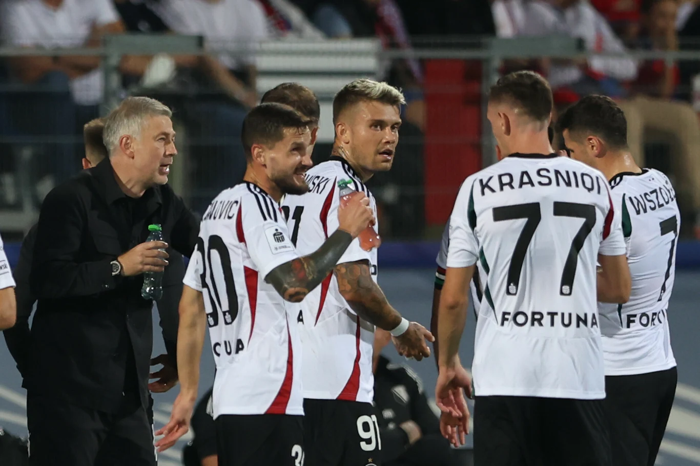 Grupa piłkarzy w biało-czarnych strojach z numerami na plecach oraz trener lub członek sztabu szkoleniowego dyskutująca podczas meczu na stadionie, na tle widocznych trybun z kibicami.