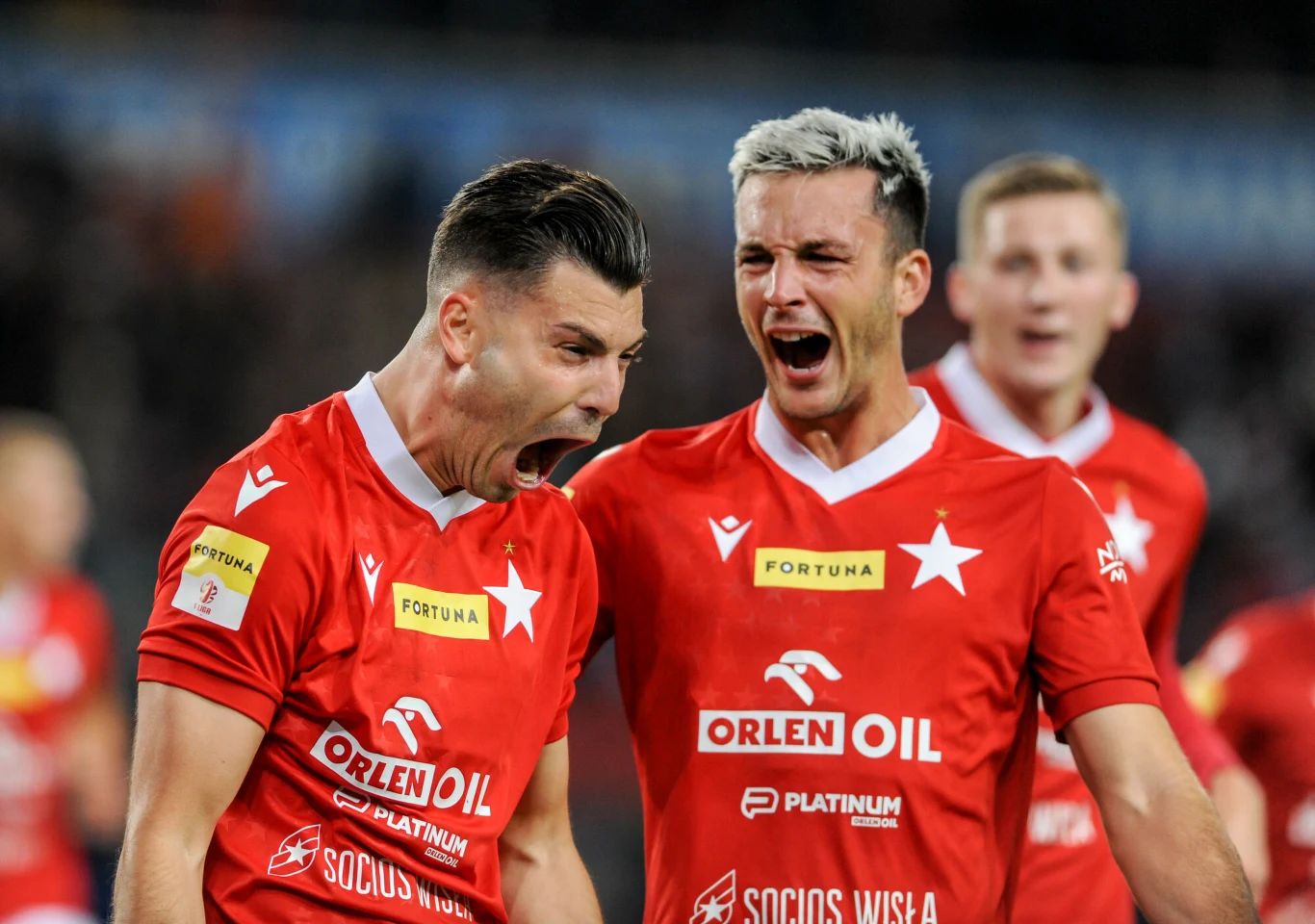 Dwaj piłkarze w czerwonych koszulkach z emblematami i logotypami sponsorów wyrażają silne emocje po zdobytym golu, jeden z nich krzyczy z radości, a drugi ma triumfalny wyraz twarzy, w tle widoczny trzeci zawodnik.