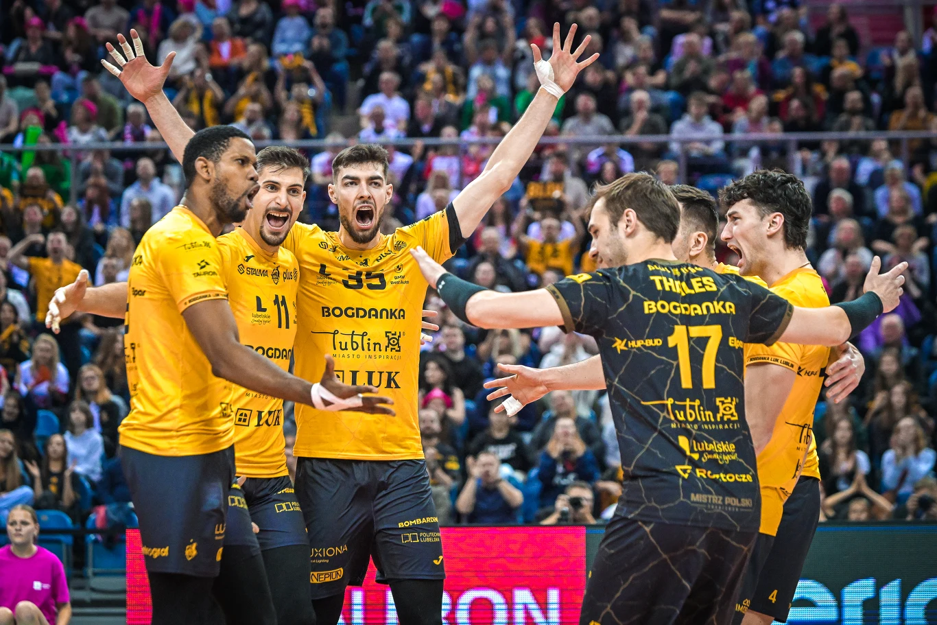 Grupa siatkarzy w żółtych strojach świętuje punkt, energicznie gestykulując i wyrażając radość, w tle widoczna publiczność w hali sportowej. Grupa siatkarzy w żółtych strojach świętuje punkt, energicznie gestykulując i wyrażając radość, w tle widoczna publiczność w hali sportowej.