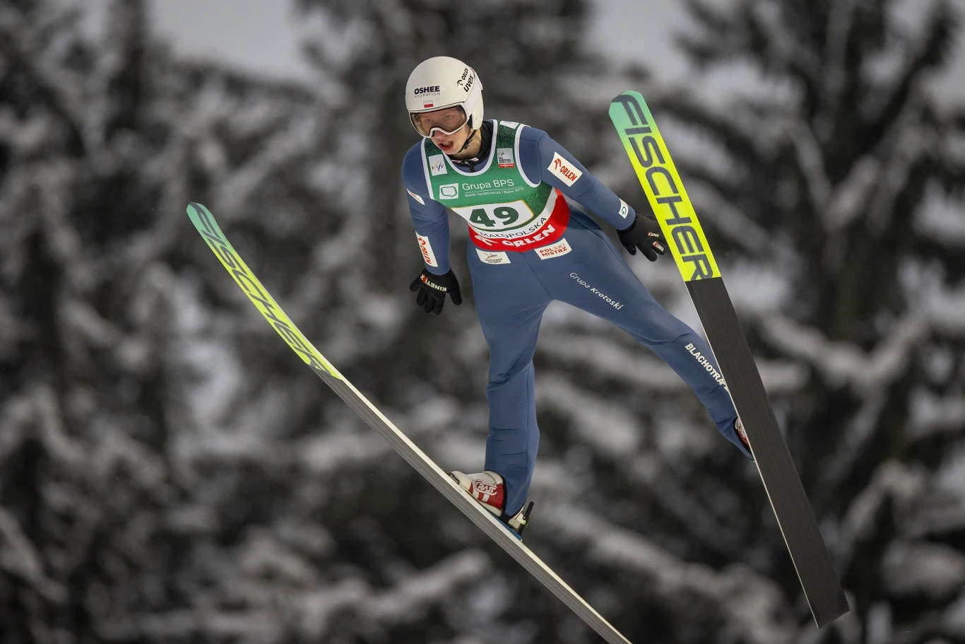 Skoczek narciarski w kombinezonie sportowym i kasku w trakcie lotu na tle rozmytych, ośnieżonych drzew, narty uniesione na boki, widoczny numer startowy 49 i logo sponsorów.