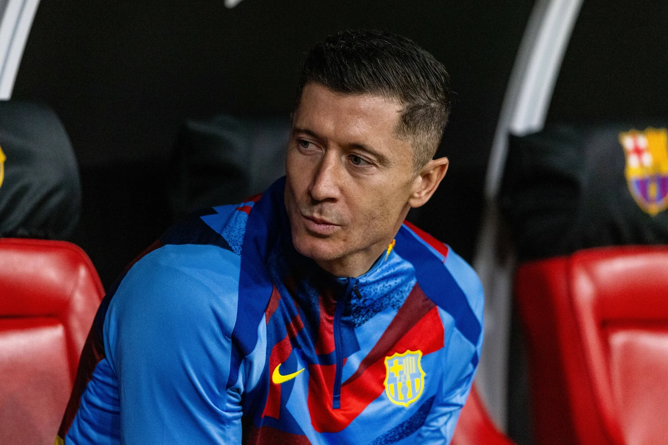 Piłkarz ubrany w niebiesko-czerwony strój sportowy z herbem klubu FC Barcelona siedzi na czerwonym fotelu, na tle widoczna część logo klubu.