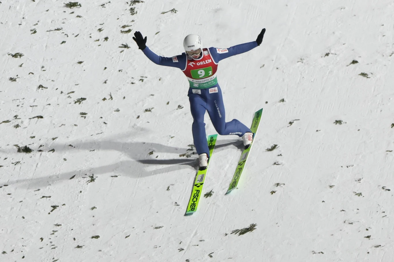 Skoczek narciarski w niebieskim kombinezonie i białym kasku ląduje na śniegu z uniesionymi ramionami, na tle białego stoku z nielicznymi kępami trawy.