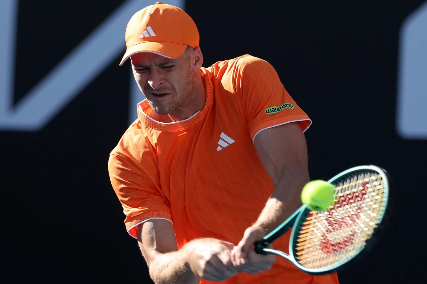 Zawodnik tenisowy w pomarańczowym stroju wykonuje uderzenie backhandowe, skupiony na piłce podczas meczu na świeżym powietrzu.