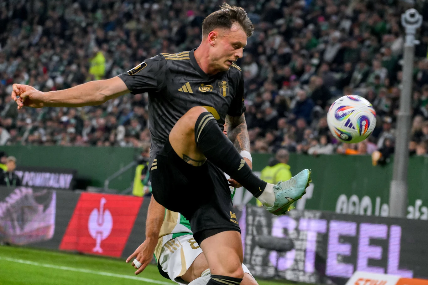 Piłkarz w czarnym stroju wykonuje dynamiczny ruch nogą podczas meczu piłki nożnej, w tle widoczna pełna trybuna kibiców oraz rozmyty zawodnik przeciwnika. Piłka w powietrzu niedaleko piłkarza, atmosfera stadionowa pełna napięcia.