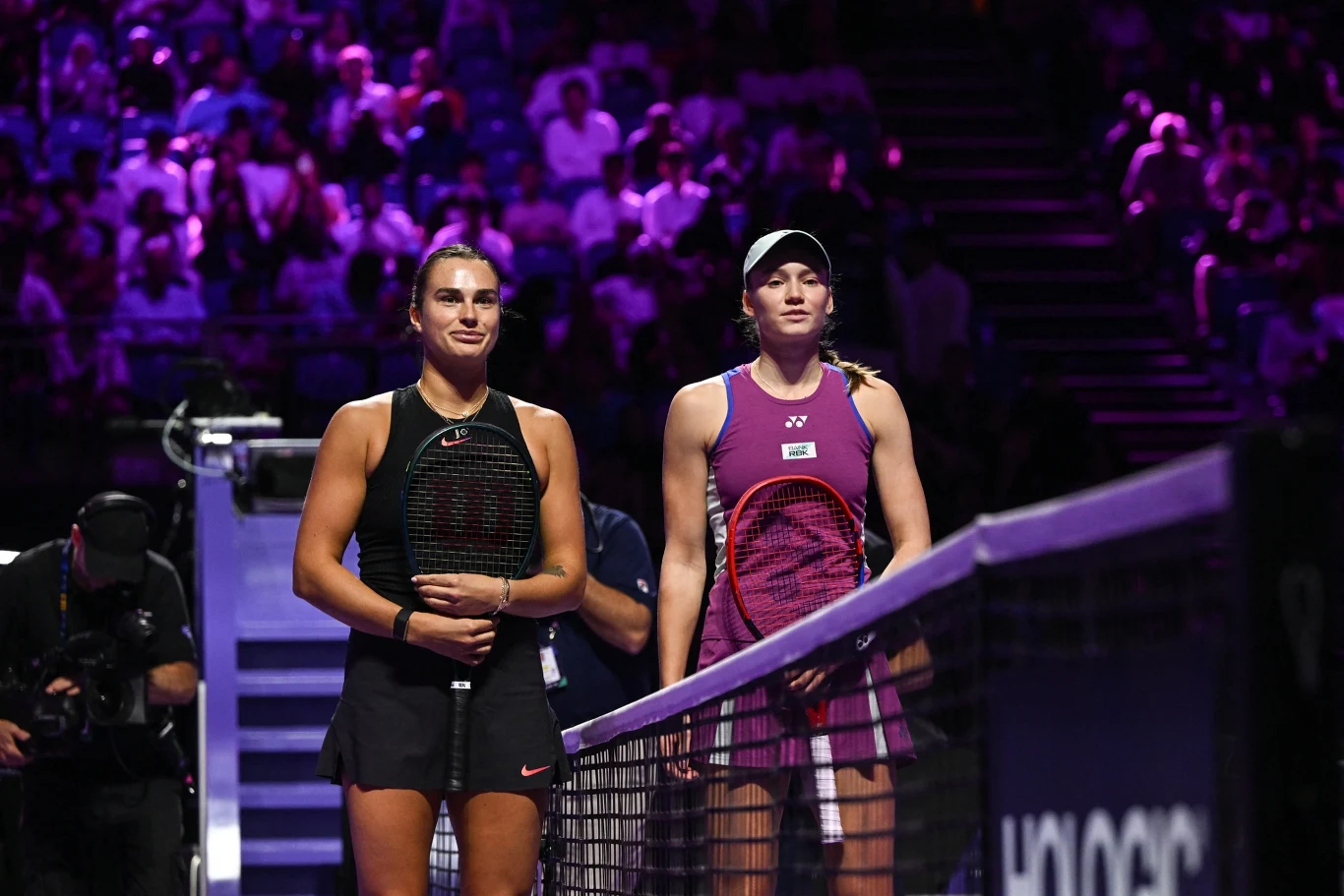Dwie zawodniczki tenisowe stoją naprzeciw siebie przy siatce na oświetlonym korcie, w tle widoczni kibice na trybunach.