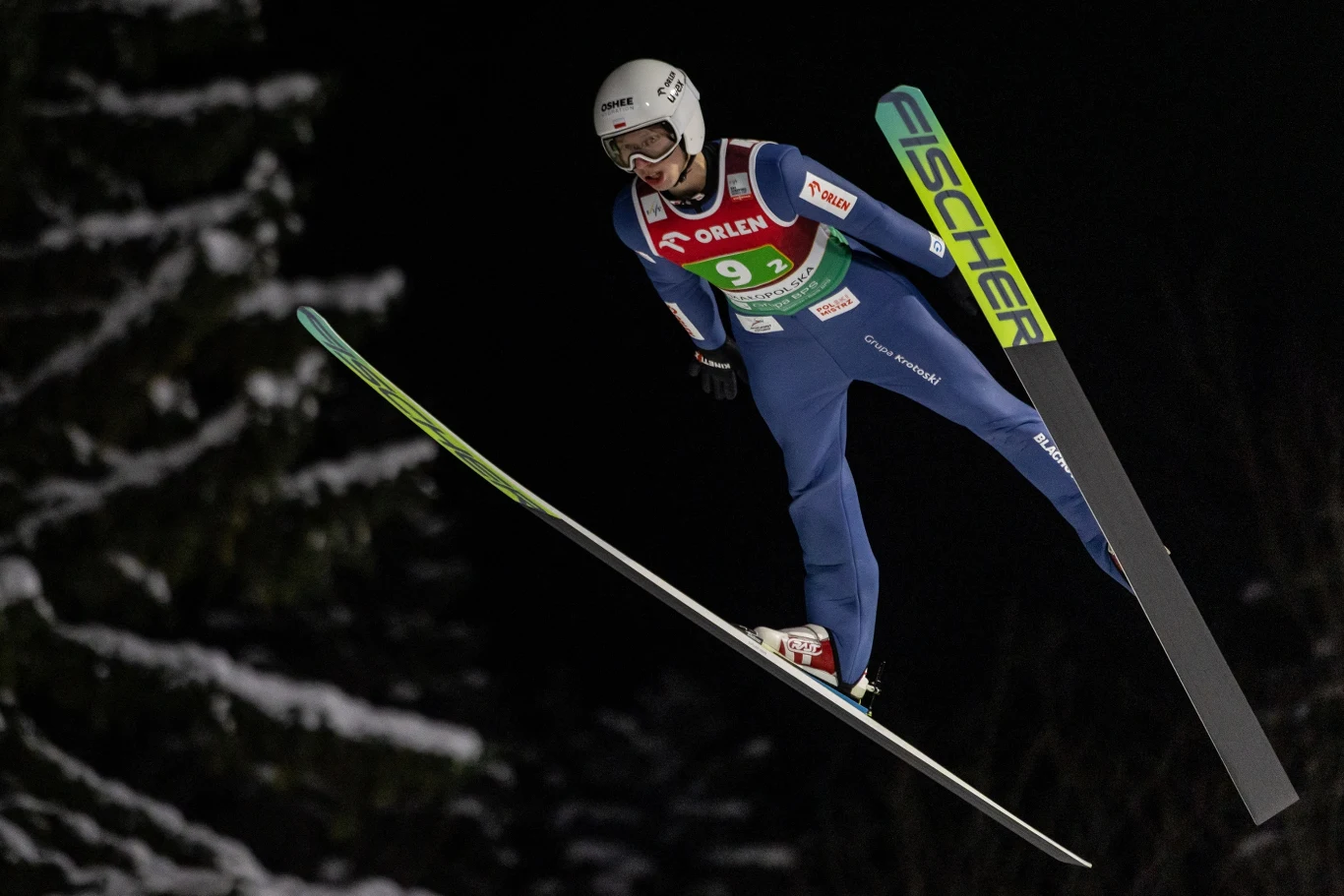 Zawodnik w stroju skoczka narciarskiego unosi się w powietrzu podczas nocnego konkursu na tle ciemnych drzew, widoczny numer startowy i sprzęt sportowy.