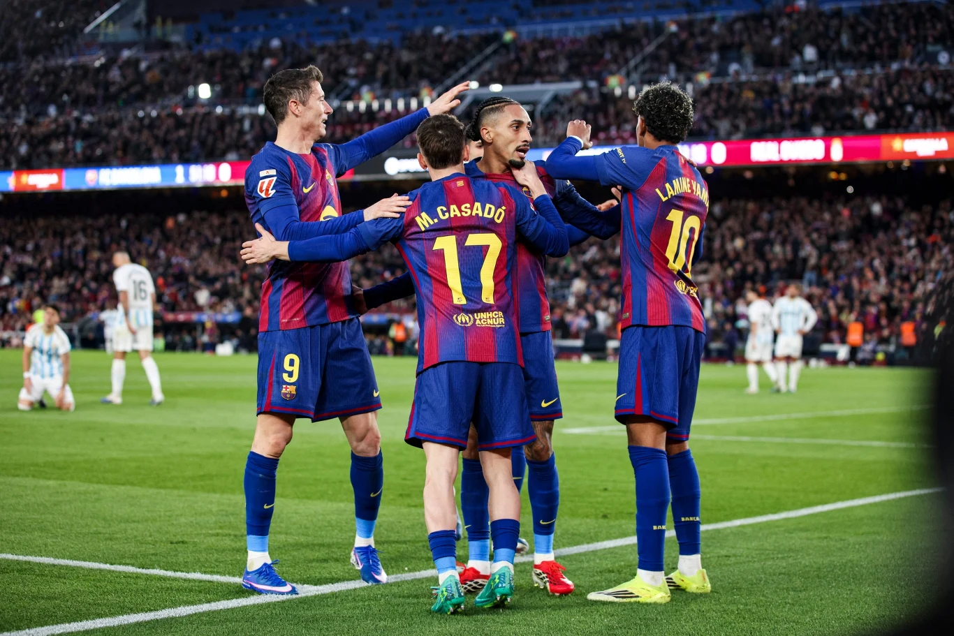 Grupa piłkarzy drużyny FC Barcelona ubranych w niebiesko-bordowe stroje świętuje wspólnie zdobycie gola na tle pełnych trybun stadionu. W tle widoczni są zawodnicy drużyny przeciwnej oraz sędzia.