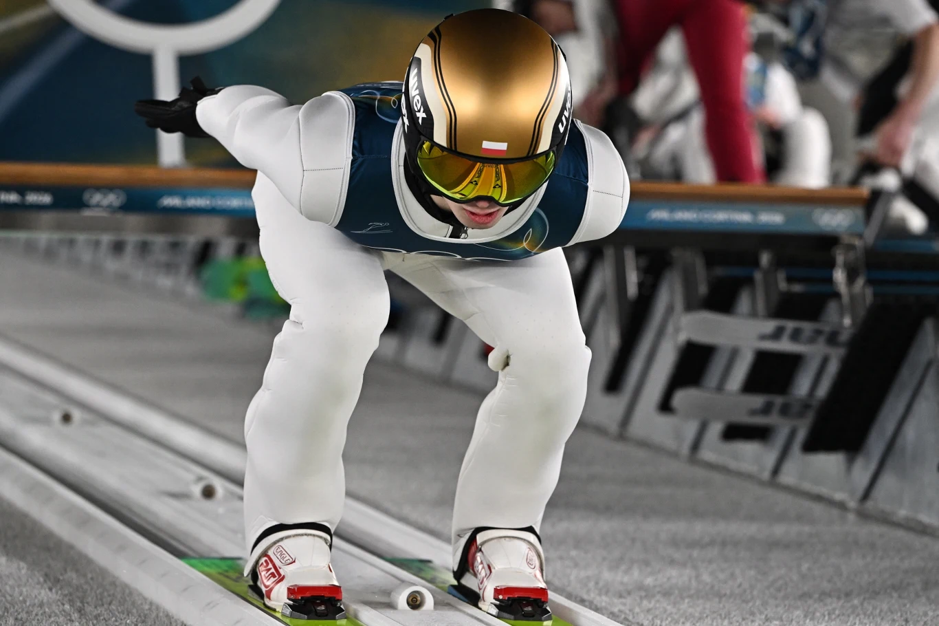 Sportowiec w kombinezonie narciarskim i kasku podczas rozpędzania się na belce startowej skoczni narciarskiej, w pozycji aerodynamicznej, gotowy do oddania skoku. Sportowiec w kombinezonie narciarskim i kasku podczas rozpędzania się na belce startowej skoczni narciarskiej, w pozycji aerodynamicznej, gotowy do oddania skoku.