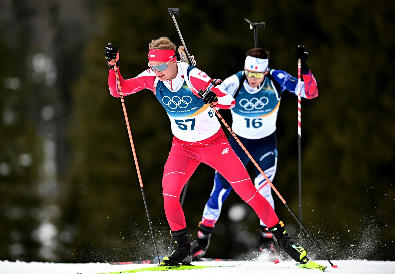 Dwójka biathlonistów podczas rywalizacji na zaśnieżonym torze olimpijskim, ubranych w sportowe kombinezony, widoczne numery startowe oraz karabiny przewieszone przez plecy.