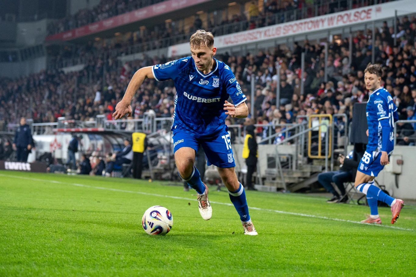 Piłkarz w niebieskim stroju drużyny Lecha Poznań biegnie z piłką przy linii bocznej boiska podczas meczu piłkarskiego na stadionie, w tle widoczni są kibice na trybunach.