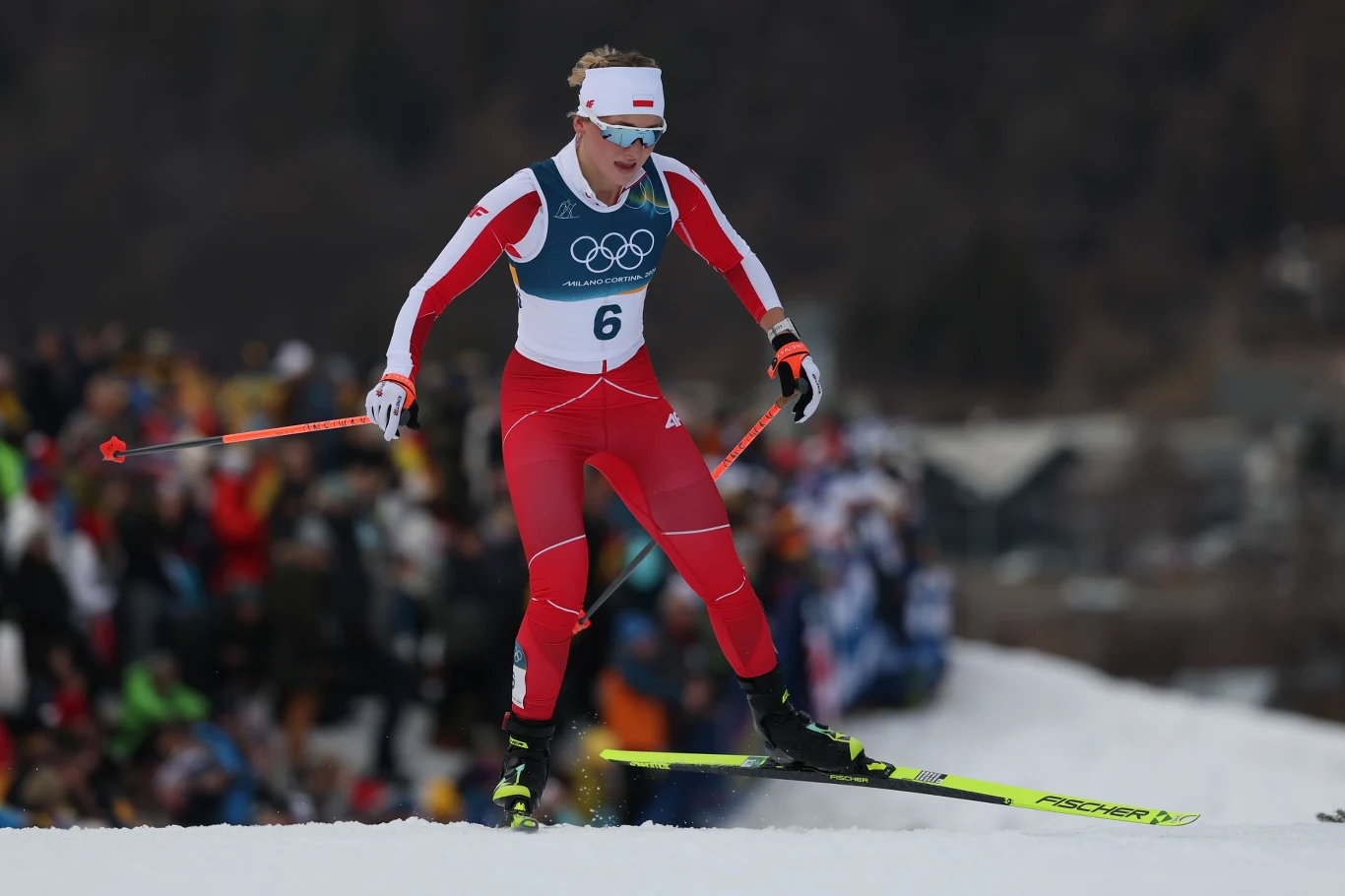 Sportowiec w czerwono-białym stroju startowym z numerem 6 biegnie na nartach biegowych podczas zawodów olimpijskich, w tle widoczny tłum kibiców.