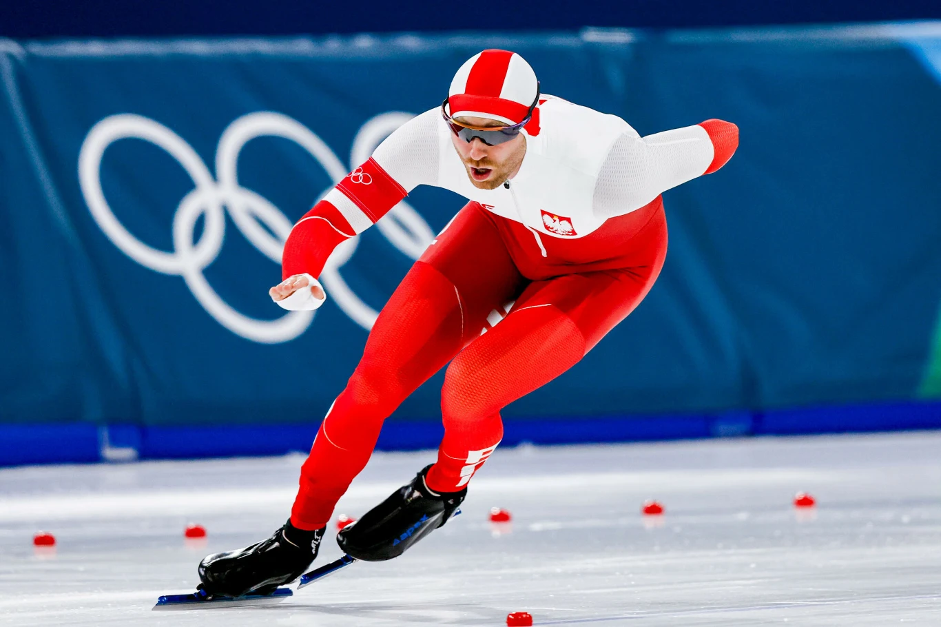 Łyżwiarz szybki w stroju reprezentacji Polski w trakcie wyścigu na lodowisku olimpijskim, wokół ustawione są znaczniki toru, w tle widoczne logo igrzysk olimpijskich.