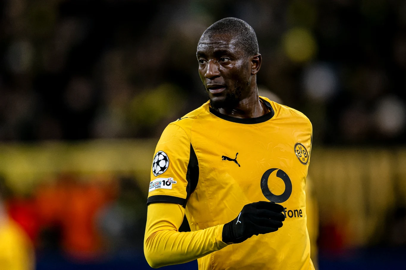 Piłkarz w żółto-czarnym stroju sportowym podczas meczu, na tle rozmytej publiczności i stadionu z wyraźnie widocznym logotypem sponsora na koszulce.