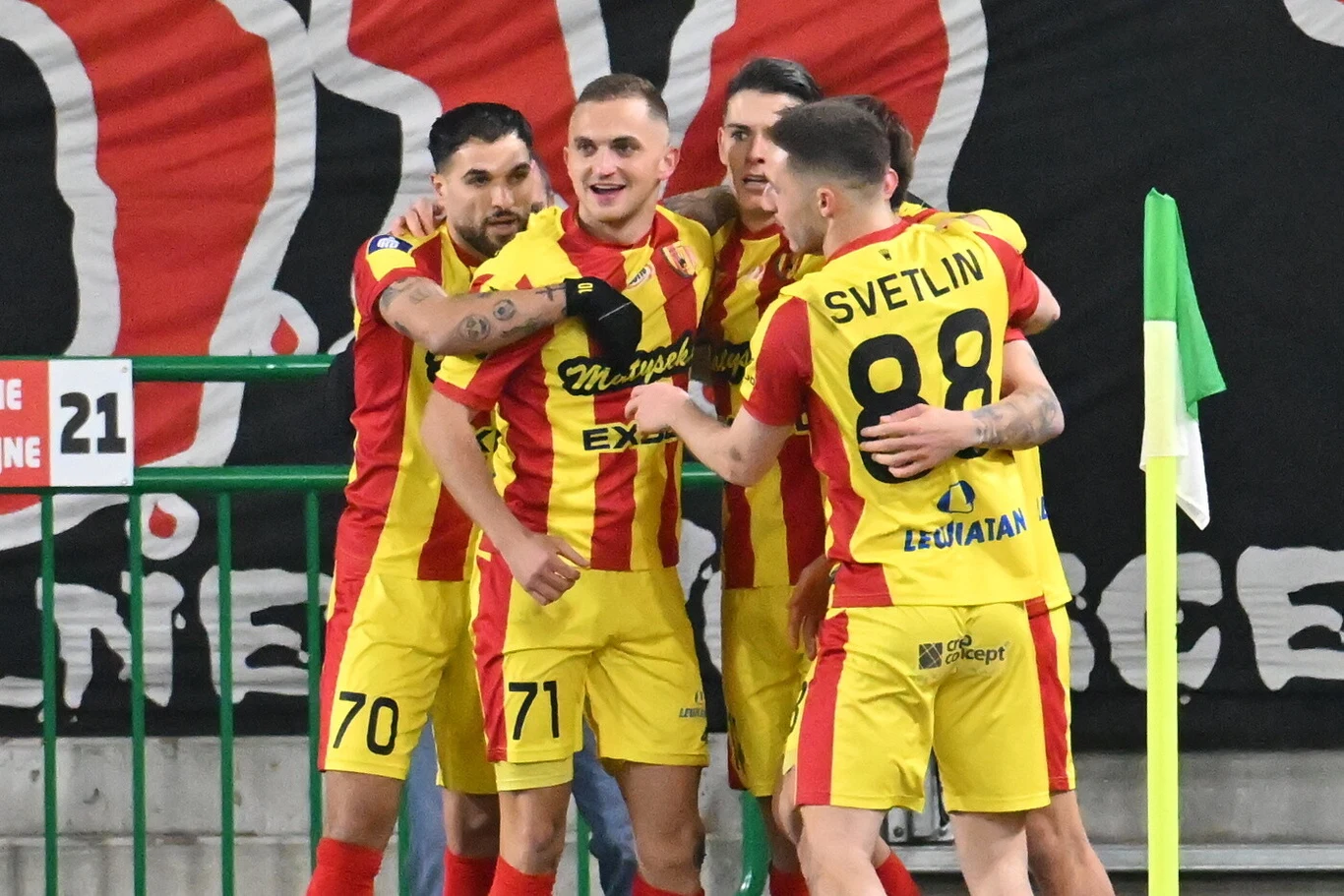 Grupa piłkarzy w żółto-czerwonych strojach świętuje zdobycie gola przy narożniku boiska, otoczona przez baner z nazwą klubu i zielone barierki.