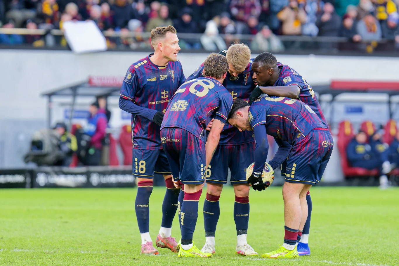 Grupa piłkarzy w granatowych strojach drużyny zebrała się w kółku podczas meczu piłkarskiego na zielonej murawie, wokół nich widoczni kibice na trybunach.