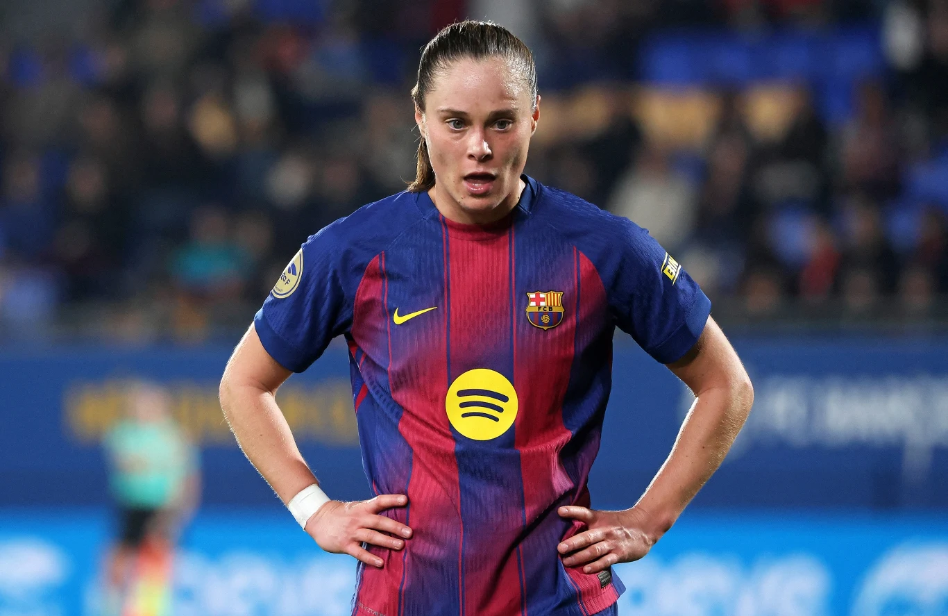 Piłkarka w stroju FC Barcelony stoi na boisku podczas meczu z rękami opartymi na biodrach, patrząc przed siebie, w tle rozmyte trybuny stadionu.