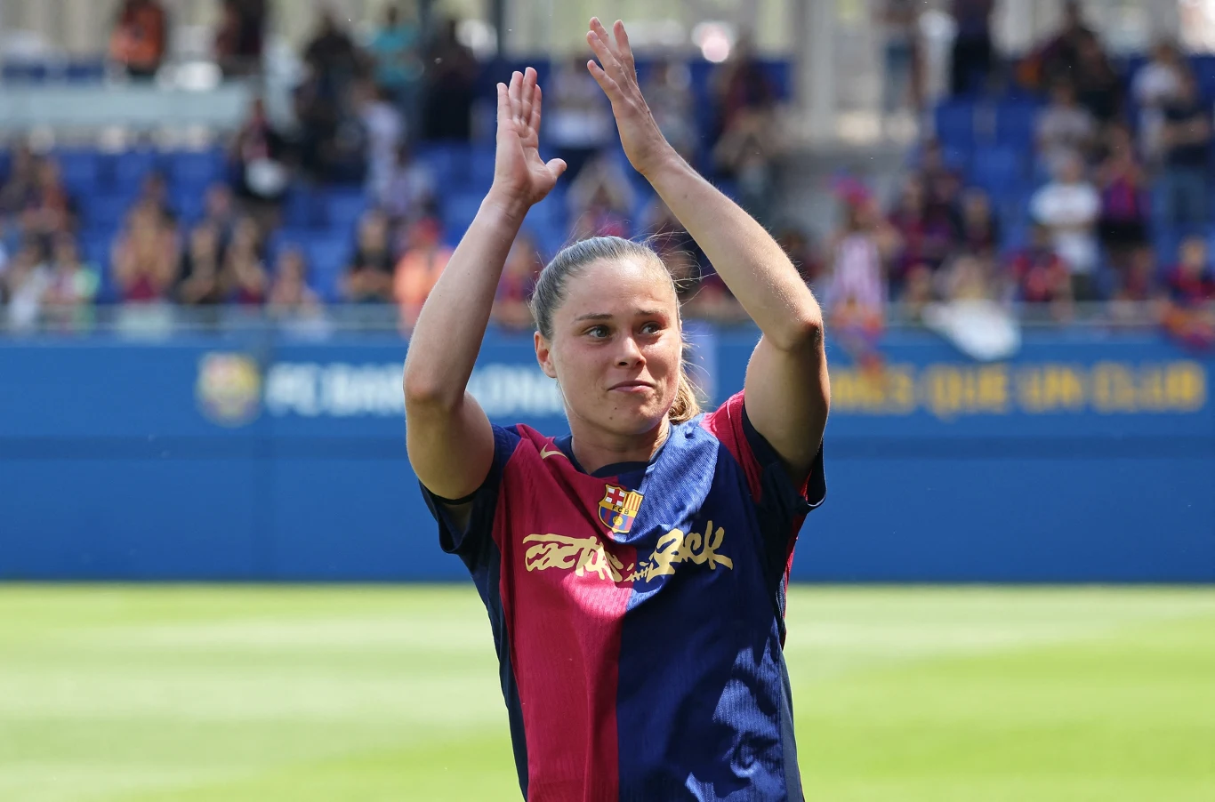 Piłkarka w stroju klubowym FC Barcelona stoi na boisku piłkarskim, unosi ręce do oklasków. W tle widoczna trybuna z kibicami oraz logo klubu. Najważniejszym elementem sceny jest zawodniczka skupiająca na sobie uwagę.